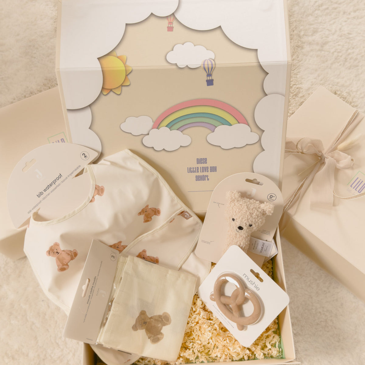 Geschenkbox für Neugeborene in Beige von lilo.littlelove mit Teddy-Rassel, Spucktüchern und Lätzchen mit Teddydesign und einem Brezelbeißring  – liebevoll verpackt in hochwertiger Erinnerungsbox mit Regenbogenmotiv