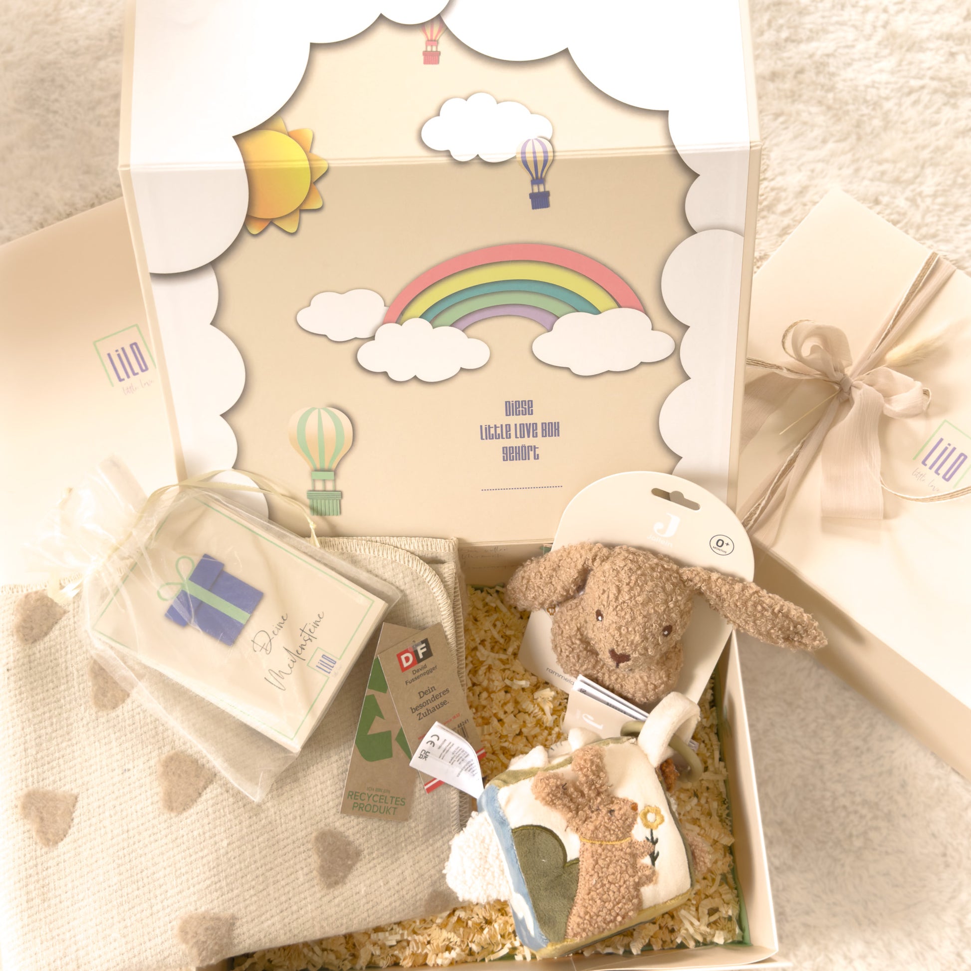 Geschenkbox für babys von lilo little love, gefüllt mit Babydecke in beige mit herzen, Meilensteinkarten, Hasenrassel aus Boucle in braun und Aktivitätenwürfel mit mehreren Motiven, u.a. ein Hase zu sehen. 